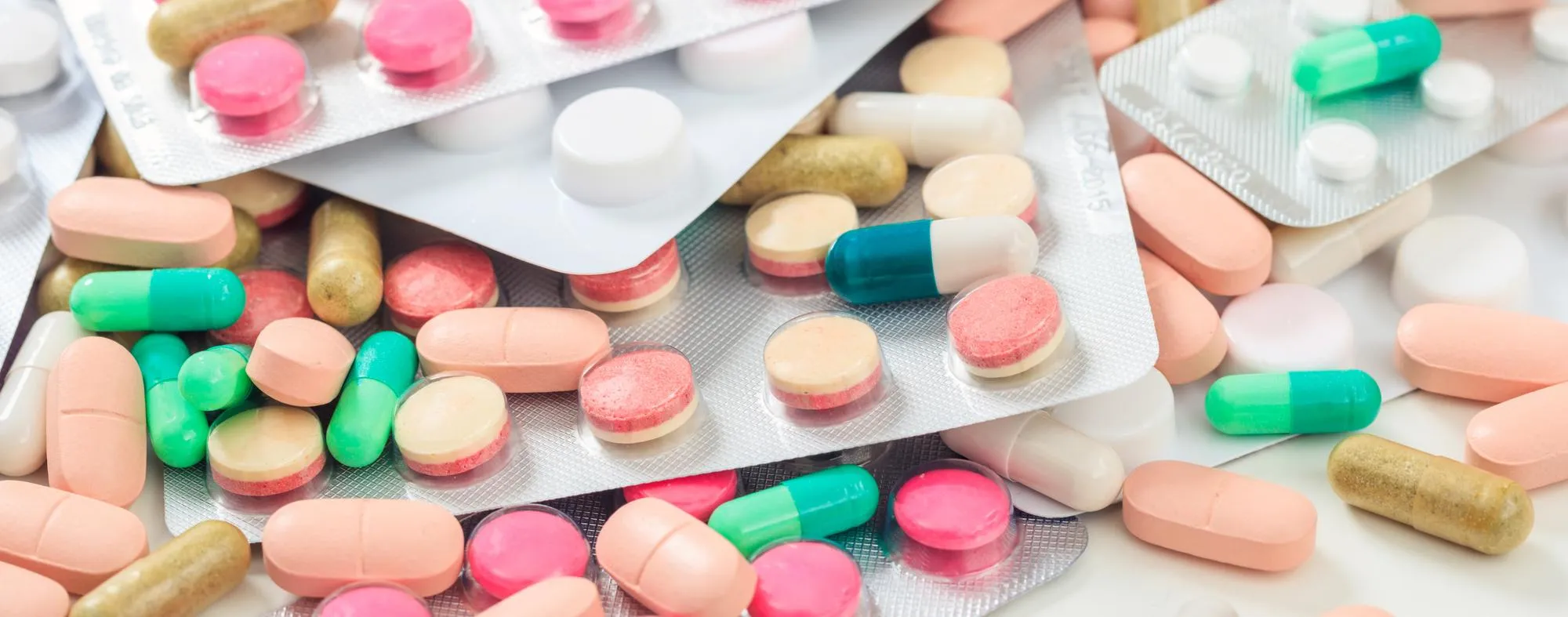 A close-up flat-lay of assorted prescription medications and over-the-counter drugs scattered on a wooden surface, including teal and yellow capsules, round tablets in white, yellow, pink, and red gel caps, alongside a silver blister pack of large round tablets highlighting the broad selection of pharmaceutical drugs and medication packaging available at a full-service pharmacy.
