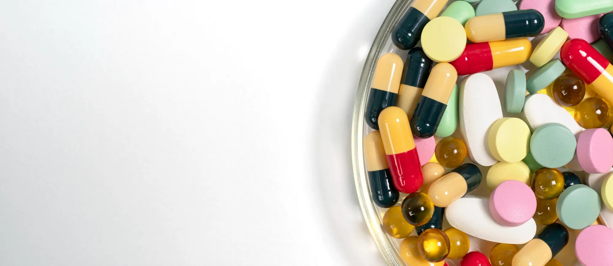 A close-up macro shot of a variety of assorted pharmaceutical tablets and capsules scattered on a wooden surface, including a clear capsule filled with white micro-beads, red-and-white capsules, a large orange-and-red capsule, round tablets in blue, pink, green, and yellow, and an oblong orange tablet representing a broad range of prescription medications, over-the-counter drugs, or pharmacy pharmaceutical offerings.