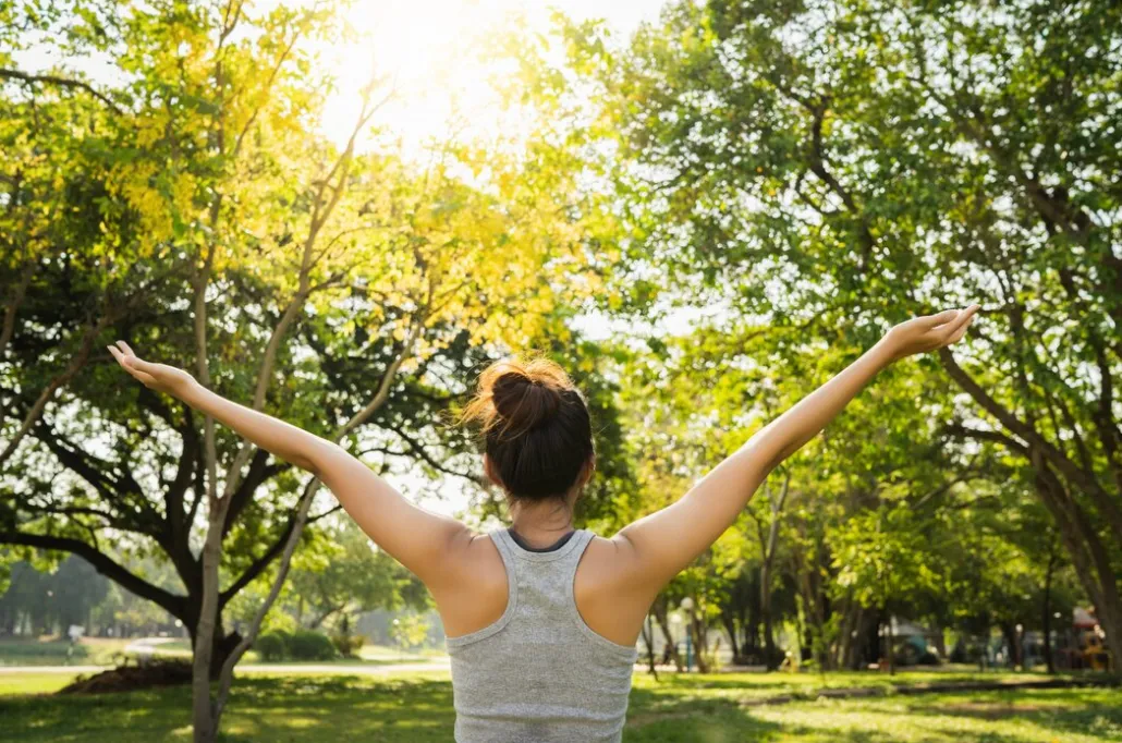 Person seen from behind with arms raised in a sunlit park, enjoying the outdoors in a green space.