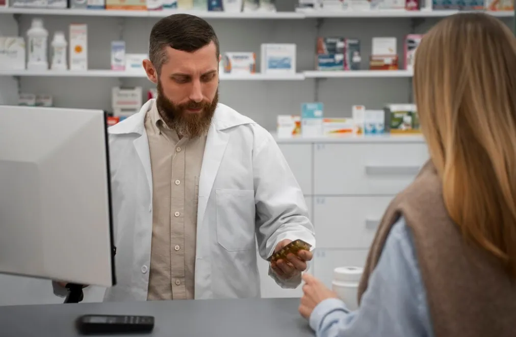 Pharmacist in a white coat hands a pill bottle to a customer at the pharmacy counter.