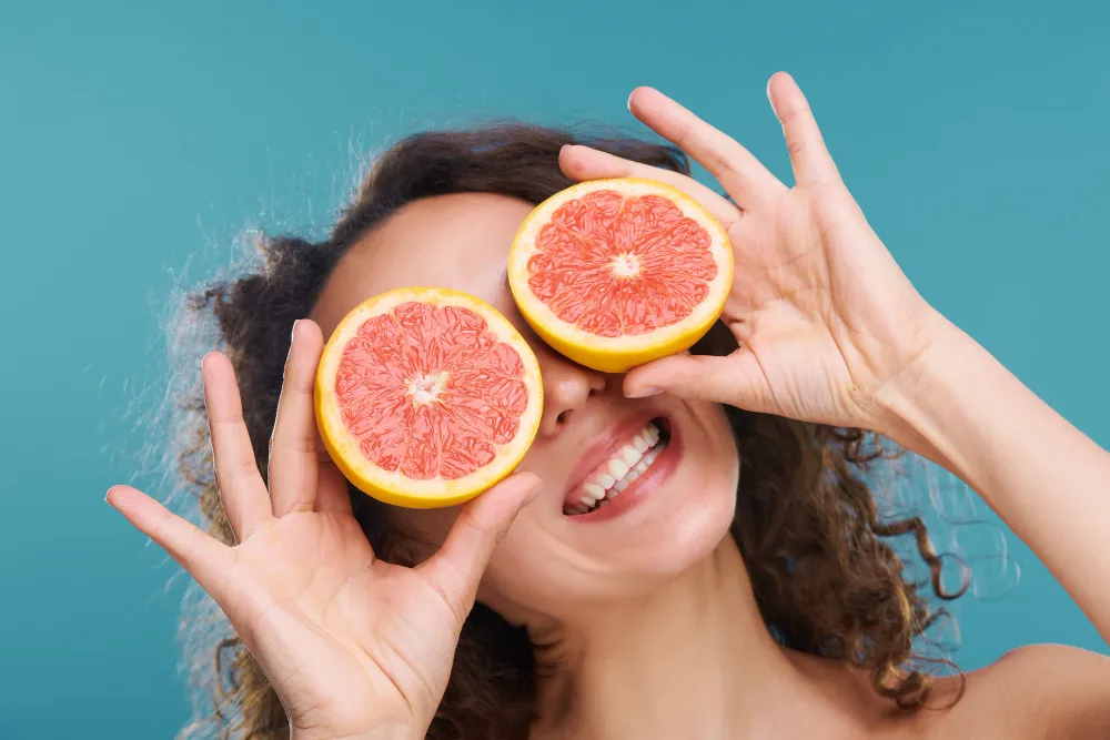 Smiling woman holding grapefruit slices over her eyes, representing a fun and healthy lifestyle supported by proper vitamin supplement intake and guidance from a trusted vitamin shoppe Jacksonville FL for balanced nutrition and multivitamins supplements