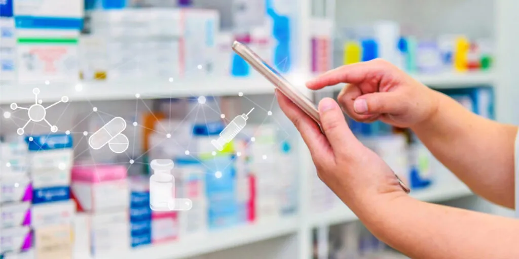 A person's hands holding and tapping a smartphone in a pharmacy, with shelves of colorful medication boxes blurred in the background, accessing an online doctor appointment platform. Overlaid on the left side of the image are glowing white digital icons connected by a network of dots and lines, depicting pharmaceutical and medical symbols including a capsule, a pill, a medicine vial, a syringe, and a molecular structure representing the concept of digital health technology, smart pharmacy, or connected healthcare.
