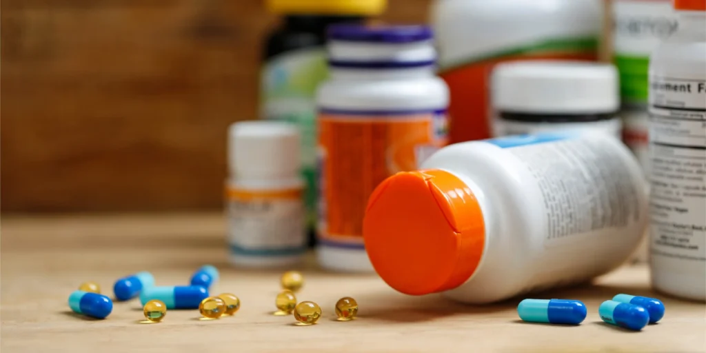 A white plastic supplement bottle with an orange cap lying on its side on a wooden surface, with assorted capsules and softgels spilled in front of it including blue capsules, teal capsules, and small golden/amber softgels, likely fish oil or vitamin E, at a vitamin and supplement store.