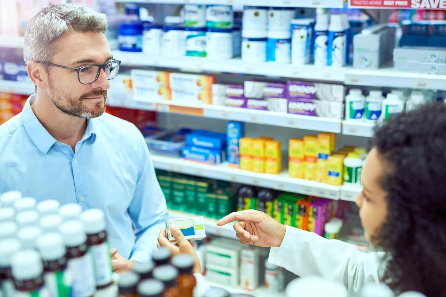 Pharmacist discussing a supplement product with a customer in a pharmacy, representing a vitamin consultation pharmacy service.