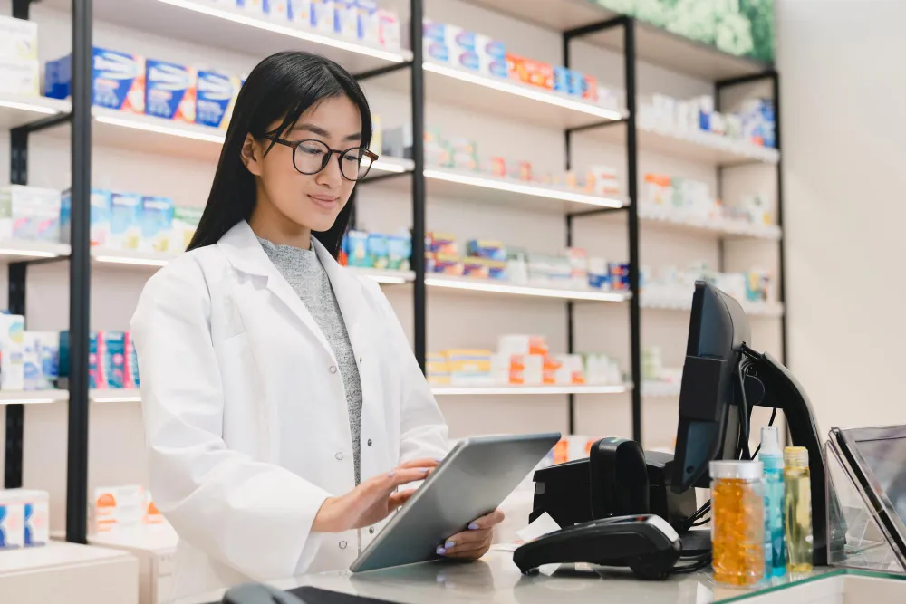 Pharmacist in a white coat using a tablet at the counter with shelves of medications behind her.